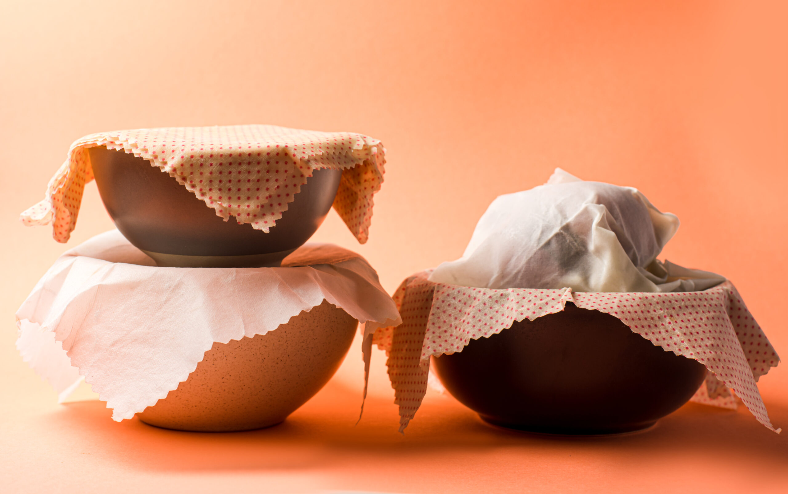 Several ceramic bowls covered with wax serviettes. Ecological type of packing or usage of wax cloth as lids for bowls. Bowls with food, isolated on light orange background.