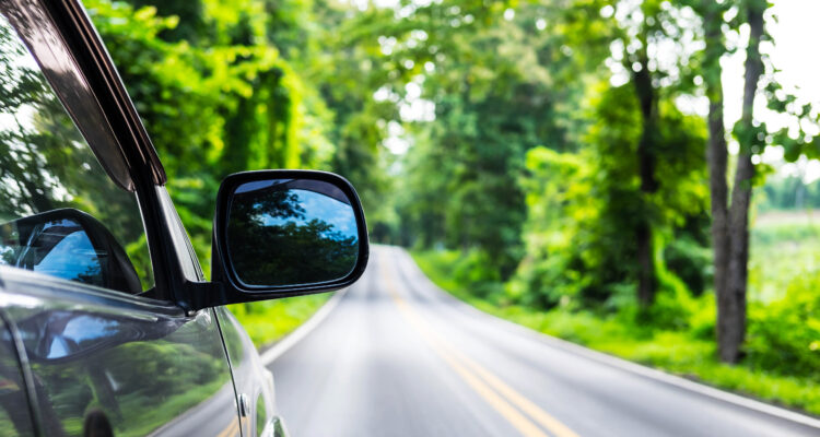 Rear view mirror of gray color car on outback road in the morning time with sunlight and blurred green forest in the background