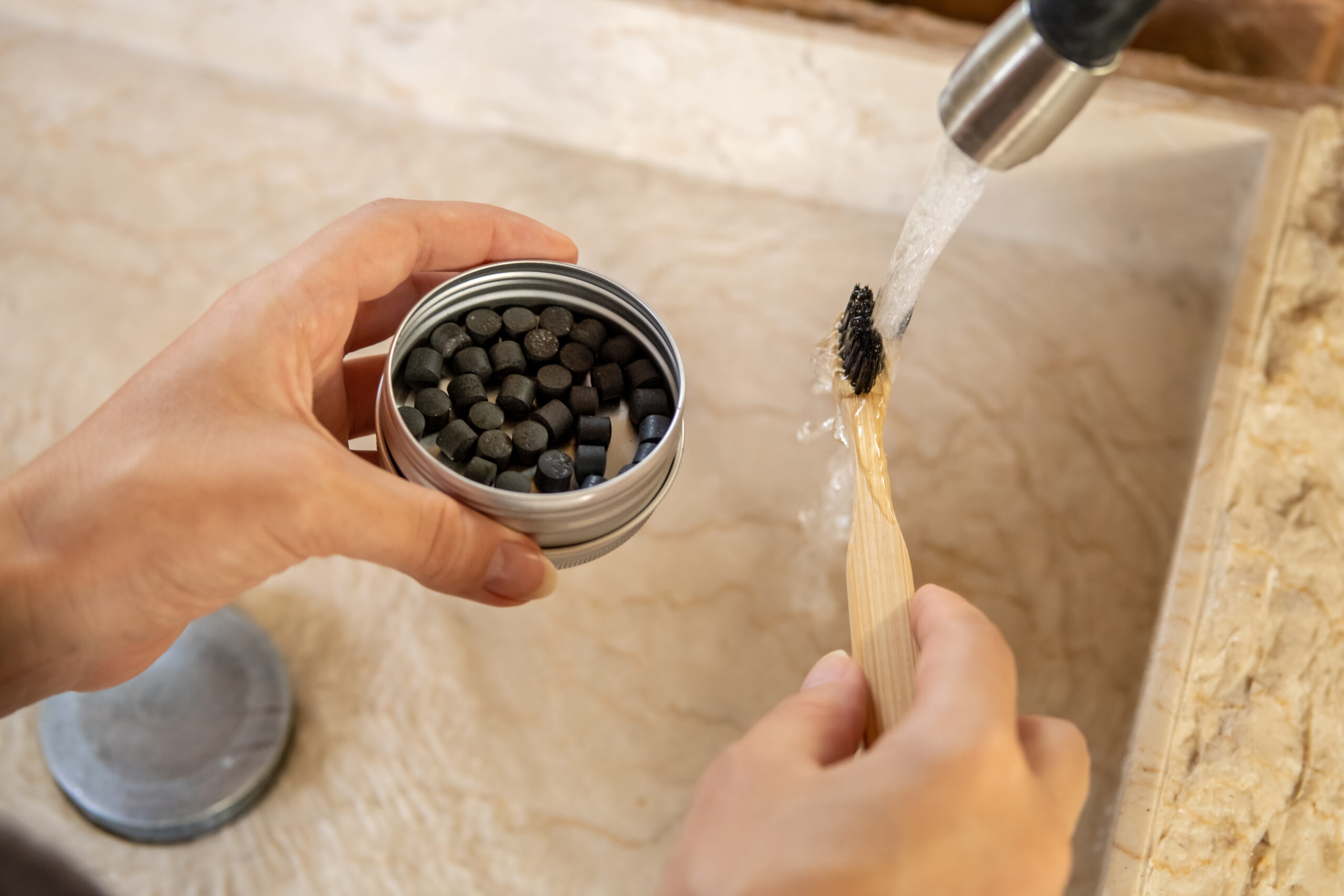 Close up of hands using eco-friendly oral care accessories by sink - biodegradable bamboo toothbrush with charcoal solid toothpaste tablets. Zero waste sustainable and plastic free lifestyle