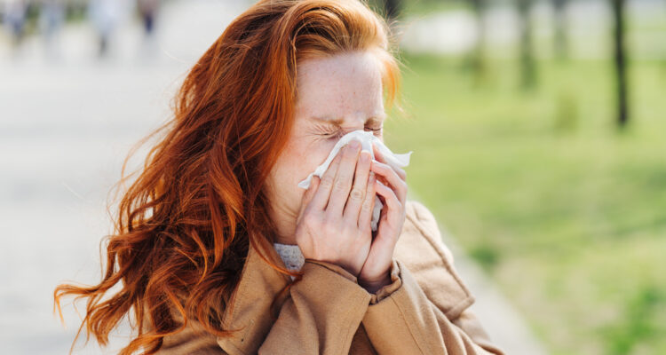 Young woman suffering from hay fever or pollen allergy in early spring blowing her nose on a tissue outdoors in a park