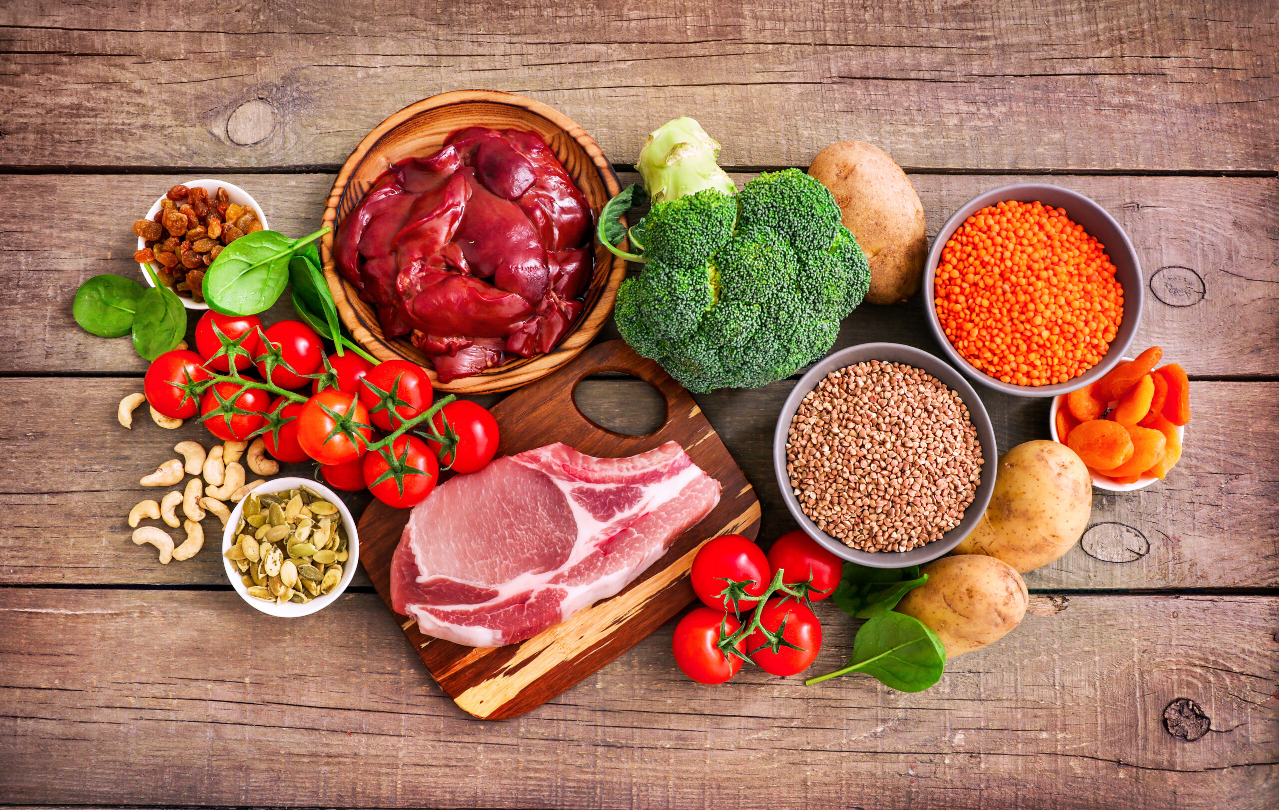 Assortment of high in Iron sources on wooden background: liver, beef, raisins, keshew, spinach, tomatoes, potatoes, buckwheat, pumpkin seeds, lentil, broccoli, dried appricots. Top view.